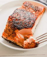 Pan-seared salmon with a crust on the top of the salmon. Set on a white plate with a wood-handled fork flaking off a large piece of moist salmon.