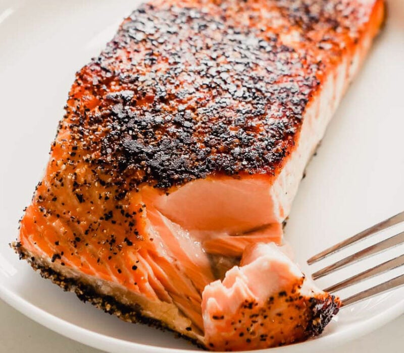 Pan-seared salmon with a crust on the top of the salmon. Set on a white plate with a wood-handled fork flaking off a large piece of moist salmon.