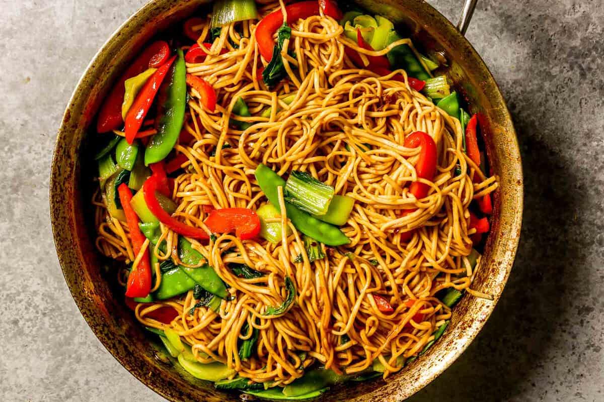 cooked lo mein noodles in a large saute pan with cooked shrimp, snow peas, bok choy and strips of red bell peppers.