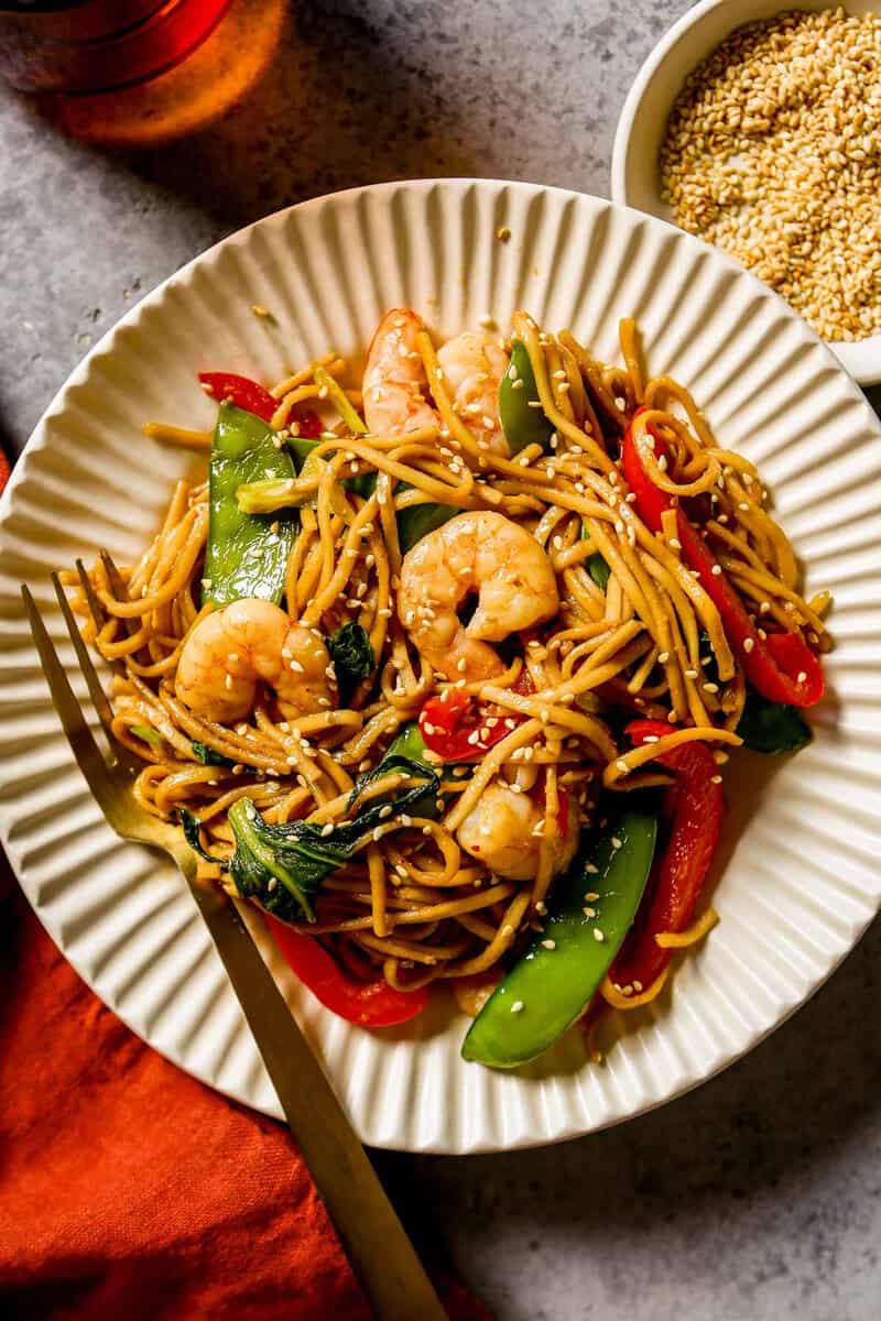 Shrimp lo mein with red bell pepper strips, snow peas, and bok choy on a ribbed plate, topped with sesame seeds. Silver fork set in the plate with a bowl of sesame seeds and a red water glass off to the side.