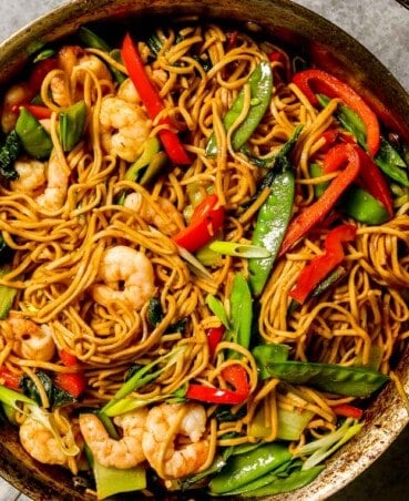 cooked lo mein noodles in a large saute pan with cooked shrimp, snow peas, bok choy and strips of red bell peppers.