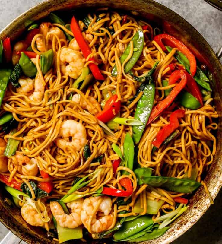 cooked lo mein noodles in a large saute pan with cooked shrimp, snow peas, bok choy and strips of red bell peppers.