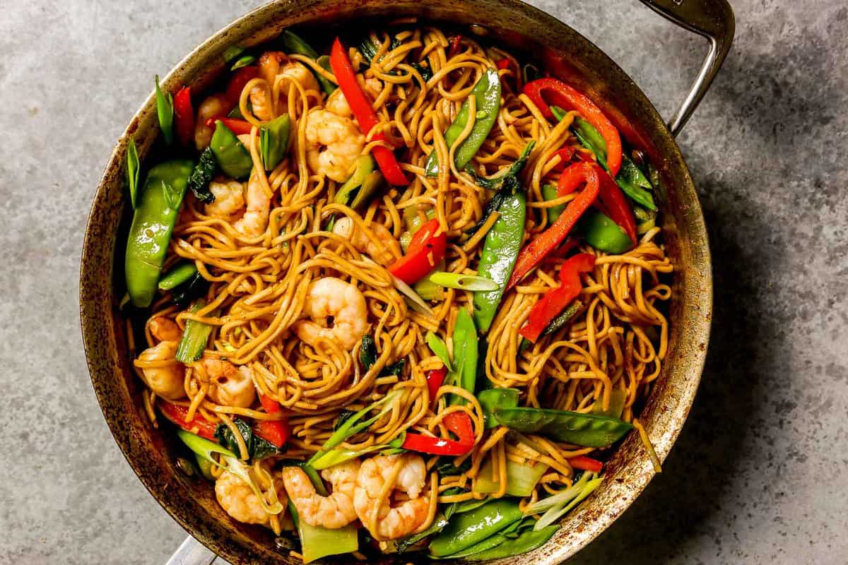 cooked lo mein noodles in a large saute pan with cooked shrimp, snow peas, bok choy and strips of red bell peppers.