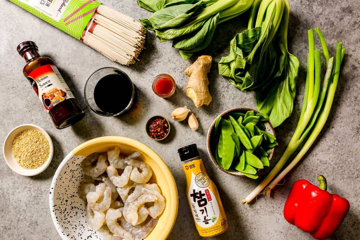 Dry lo mein noodles, fresh bok choy, scallions, hoison sauce, soy sauce, red pepper flakes, sriracha, fresh ginger, garlic cloves, fresh snow peas, red bell pepper, sesame seeds and raw peeled and deveined shrimp set out on a counter. 