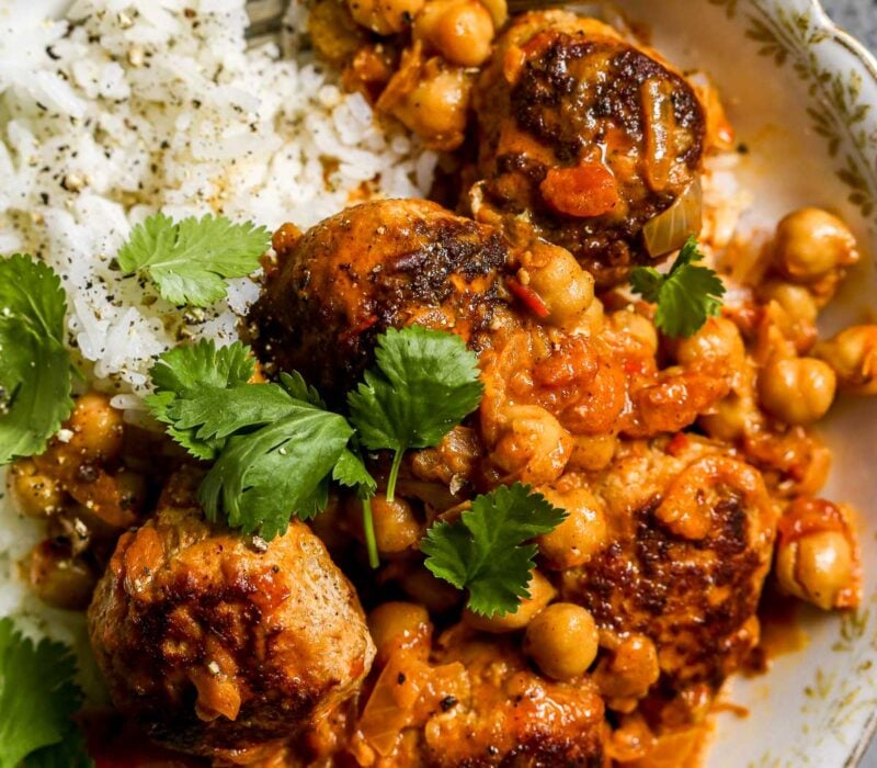 Gold and white bowl filled with rice and topped with meatballs and chickpeas coated in a creamy harissa sauce. Topped with a few cilantro leaves.