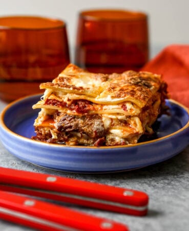 Slice of lasagna al on a small blue plate. Chunks of shredded been, melty cheese and tomato sauce oozing out of the layers of lasagna noodles.