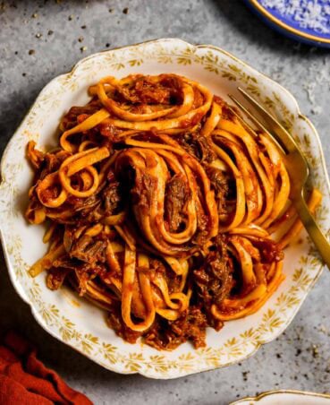 Linguine coated in a sauce with chunks of shredded short rib throughout, swirled into white bowl with gold etching on the edges. A gold fork is set in the bowl with the pasta.