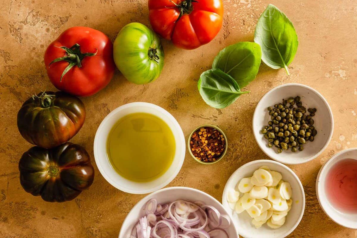 heirloom tomatoes, olive oil, sliced shallots, sliced garlic, red wine vinegar, capers, red pepper flakes and basil arranged on a counter