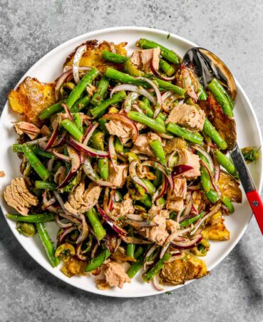 Potato salad with tuna and green beans on a large plate along with slices of red onion and olives.