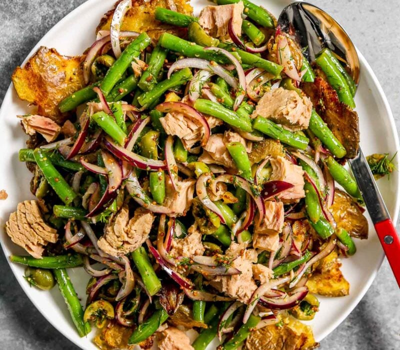 Potato salad with tuna and green beans on a large plate along with slices of red onion and olives.