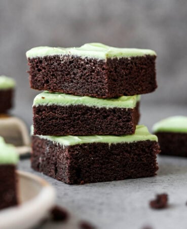 A stack of three slices of chocolate cake, each slice topped with a layer of green mint frosting. Chocolate cake crumbs scattered in front of the stack of cake slices.