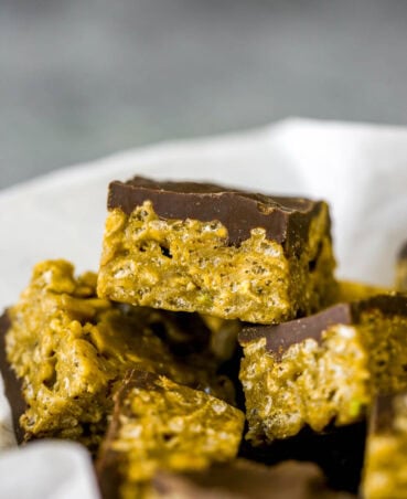 Small pistachio bars stacked on top of eachother in a bowl lined with white parchment paper. Each green pistachio bar is covered with a layer of chocolate on top.