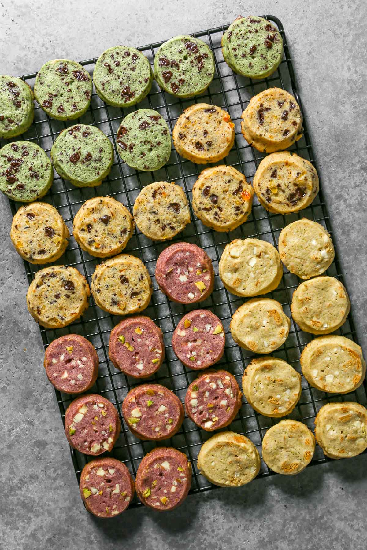 Icebox cookies baked and cooling on a wire rack. All the cookies are different flavors and colors including green cookies, pink cookies and cookies with chocolate chunks in them.