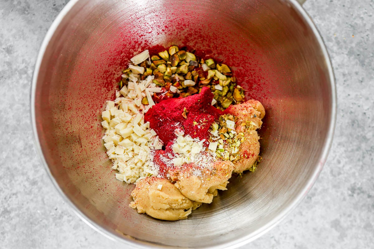Cookie dough in a metal mixing bowl topped with pistachios, white chocolate and raspberry powder.
