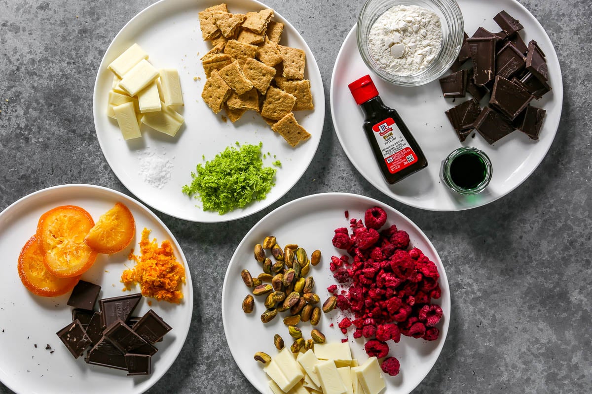 Four plates with cookie mix-ins set on each include a plate with graham crackers, lime, and white chocolate, a second one with orange and dark chocolate, a third with pistachios, freeze dried raspberries, and white chocolate, and the last one with dark chocolate, flour, mint extract and creme de menthe in a glass jar. 