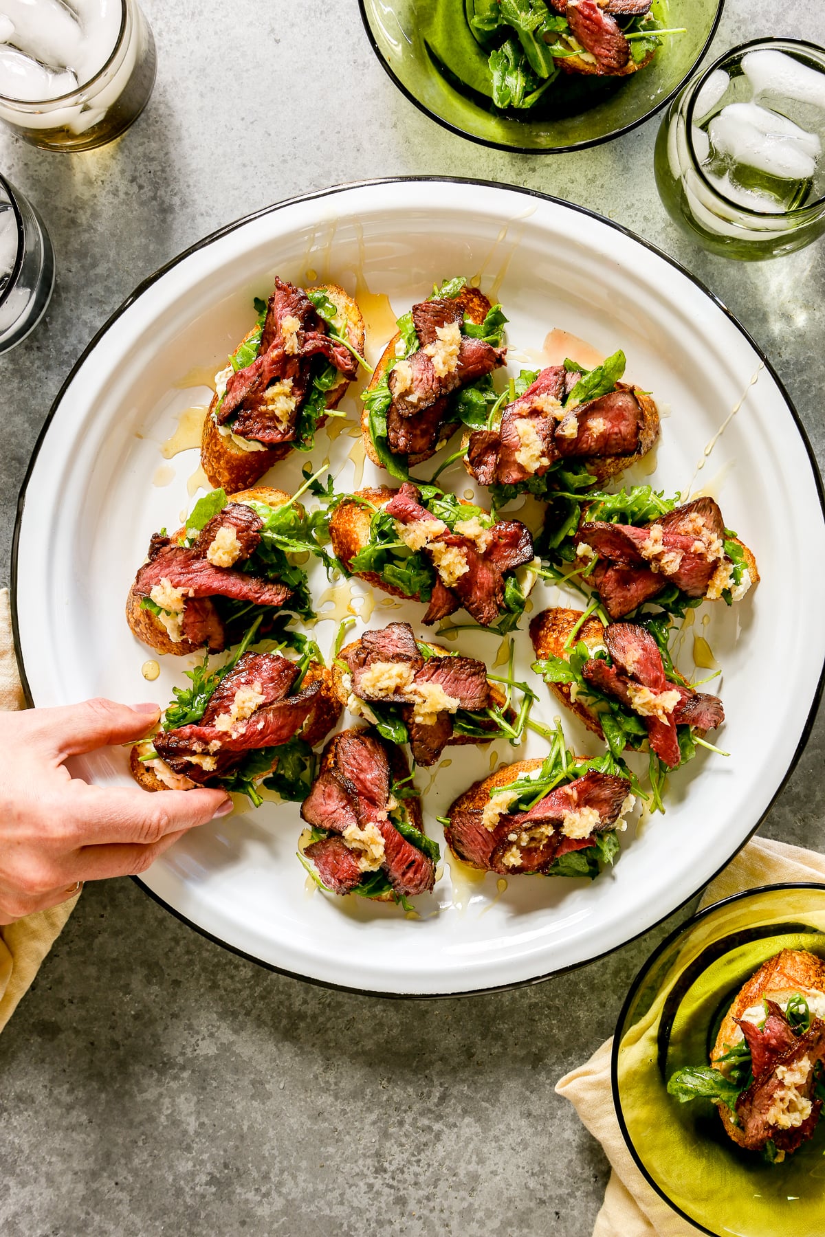 Steak crostini arranged on a large white platter with someone picking one off the plate. Water glasses and appetizer plates set around.