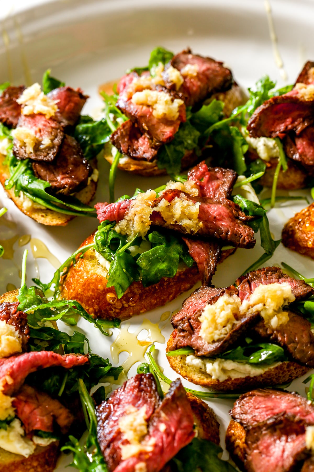 Crostini topped with sliced steak, arugula, cheese, and a drizzle of sauce on a white plate.