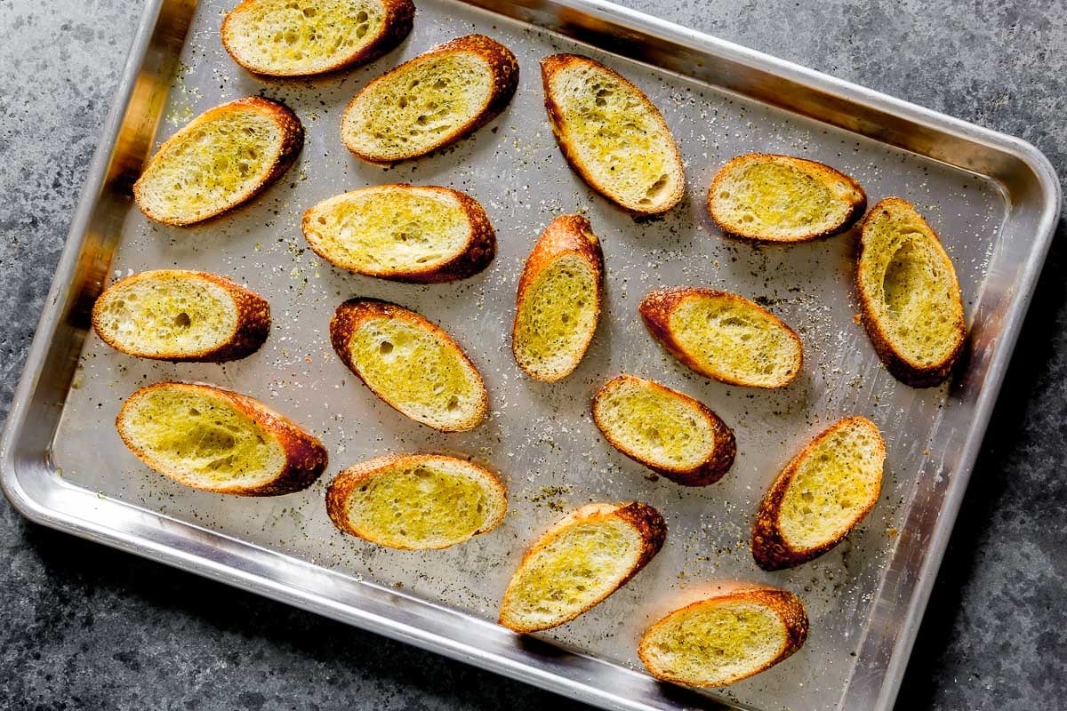 Sliced toasted baguette pieces on a metal baking sheet, lightly seasoned with salt and pepper.