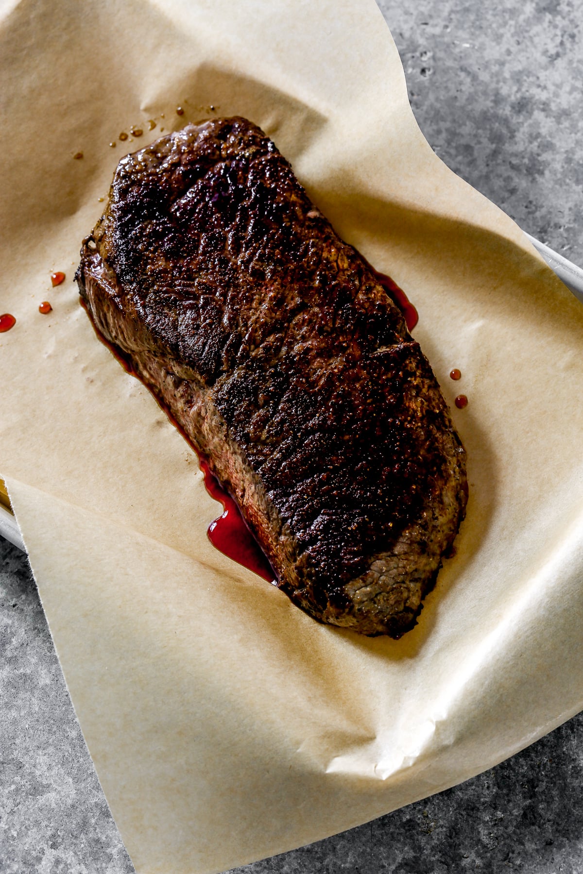 A cooked steak for steak crostini rests on brown parchment paper with visible meat juices on a gray surface.