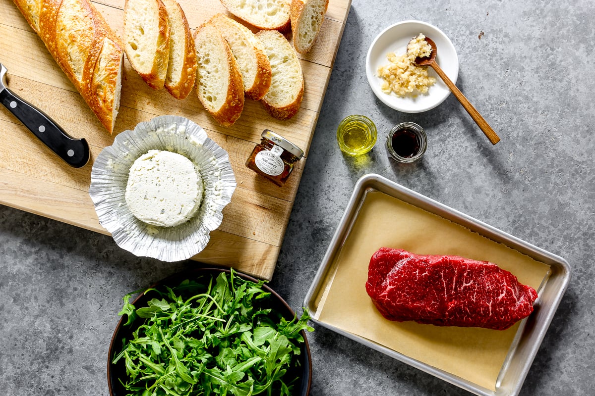 Sliced baguette, steak, arugula, Boursin cheese, olive oil, red wine vinegar, horseradish, and honey set out on a counter.