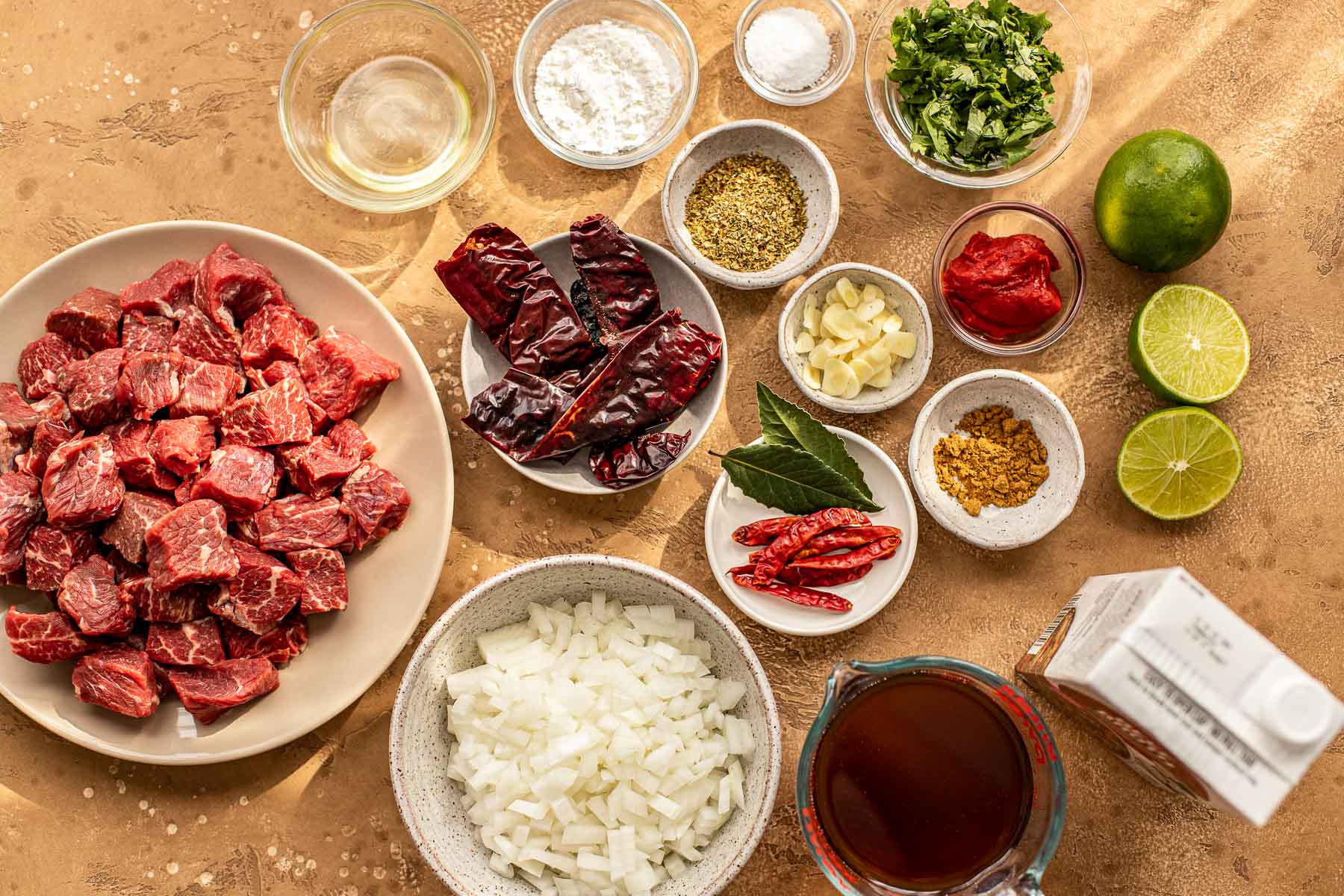 Various bowls with cubed beef, dried chilies, chopped onion, dried oregano, cumin, sliced garlic, tomato paste, cornstarch, bay leaves, lime and beef broth.
