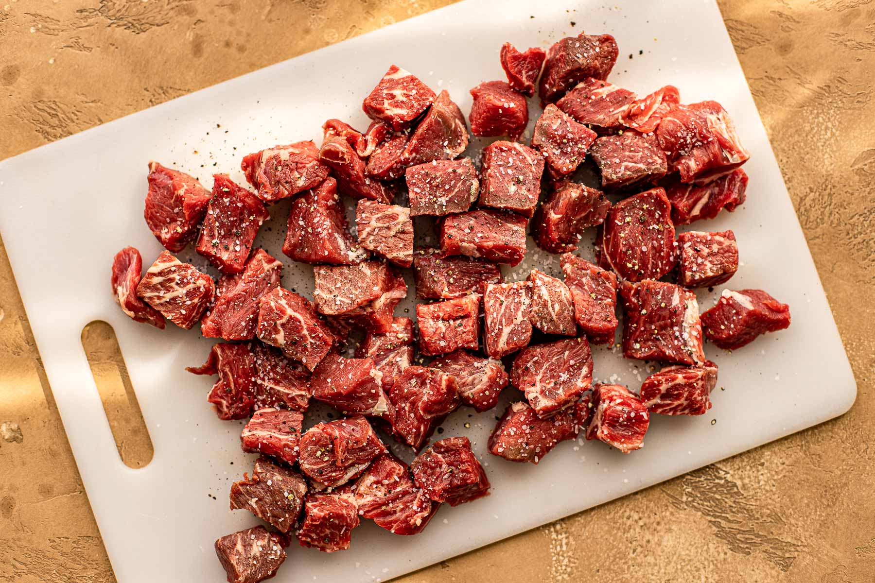 Cubed pieces of raw chuck roast with seasoning on a white cutting board, set on a textured tan surface.