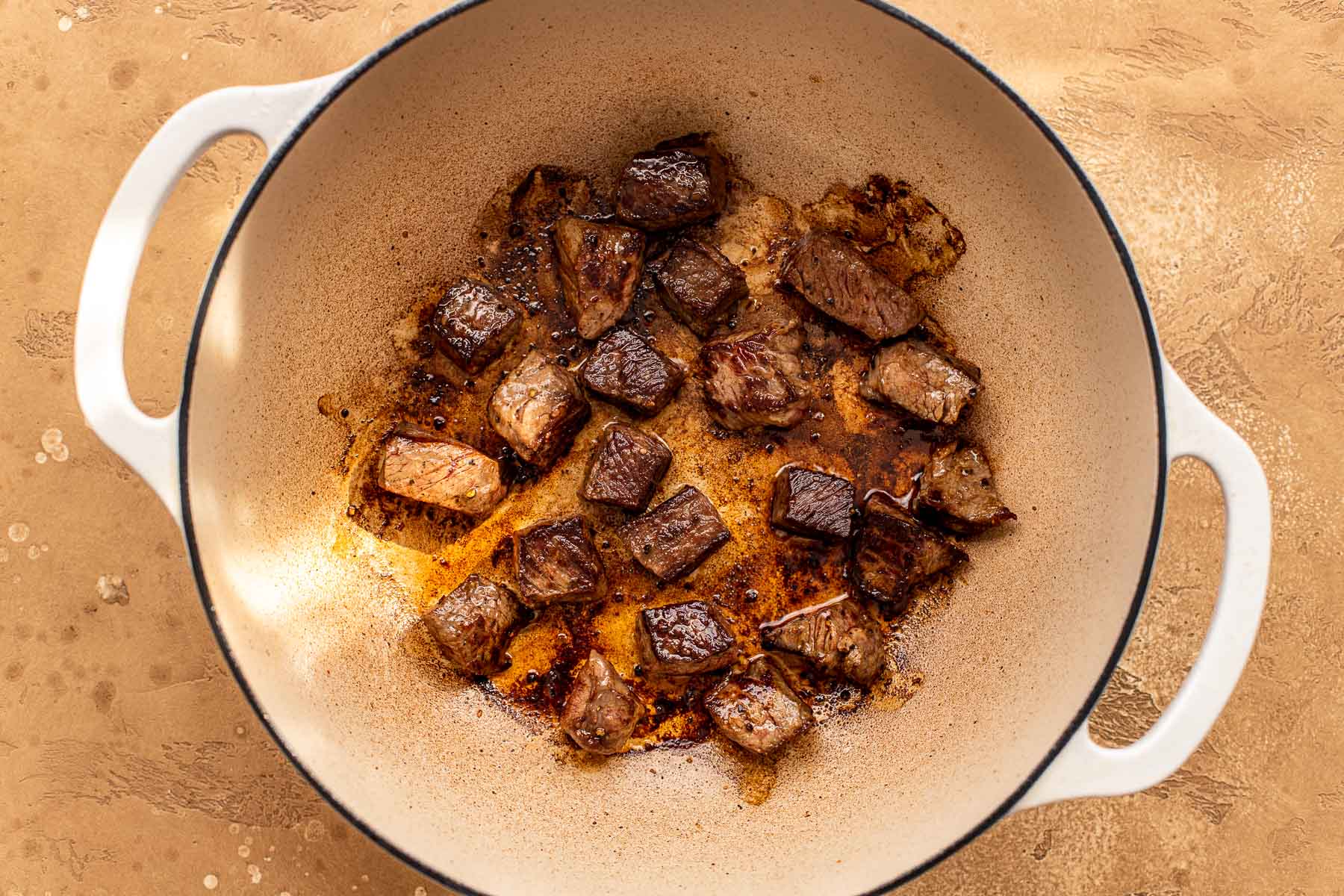 Cubed pieces of browned chuck roast cooking in a white Dutch oven on a tan countertop.