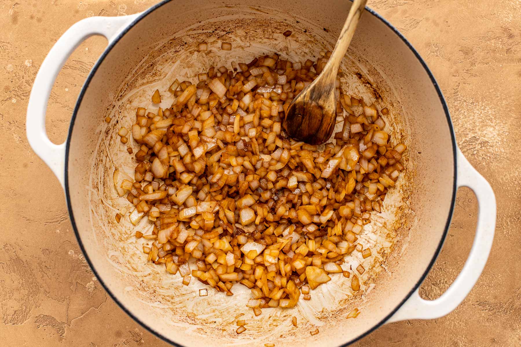 Diced onions being sautéed in a large white pot, stirred with a wooden spoon on a tan surface.