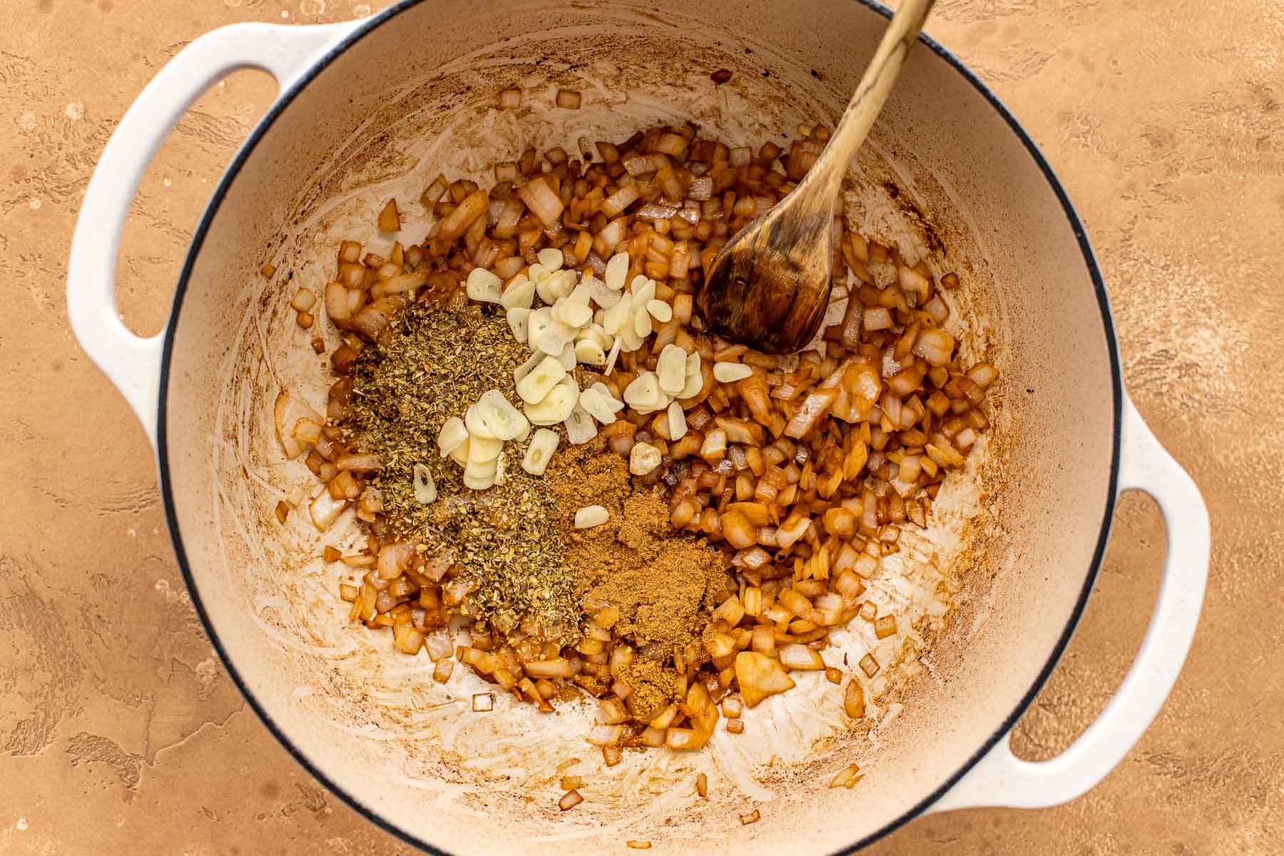 Chopped onions, sliced garlic, dried oregano and cumin cooking in a white pot with a wooden spoon on a tan surface.