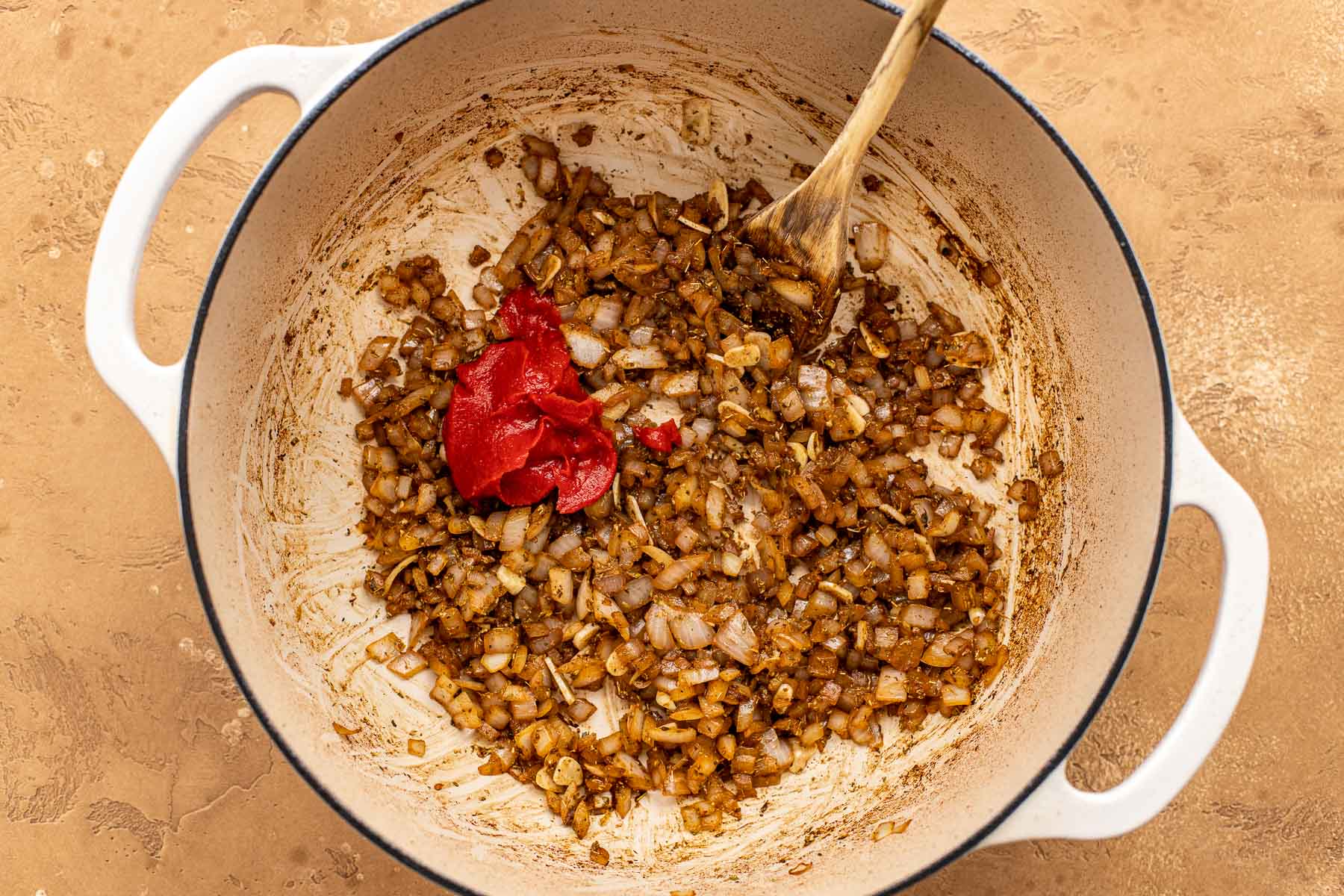 Diced onions and spices sautéing in a large white pot, with tomato paste added and a wooden spoon resting inside.