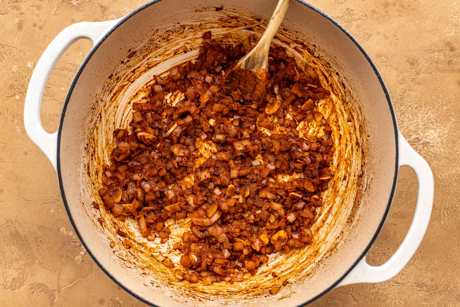 Chopped onions, spices and tomato paste being sautéed in a large white pot with a wooden spoon.