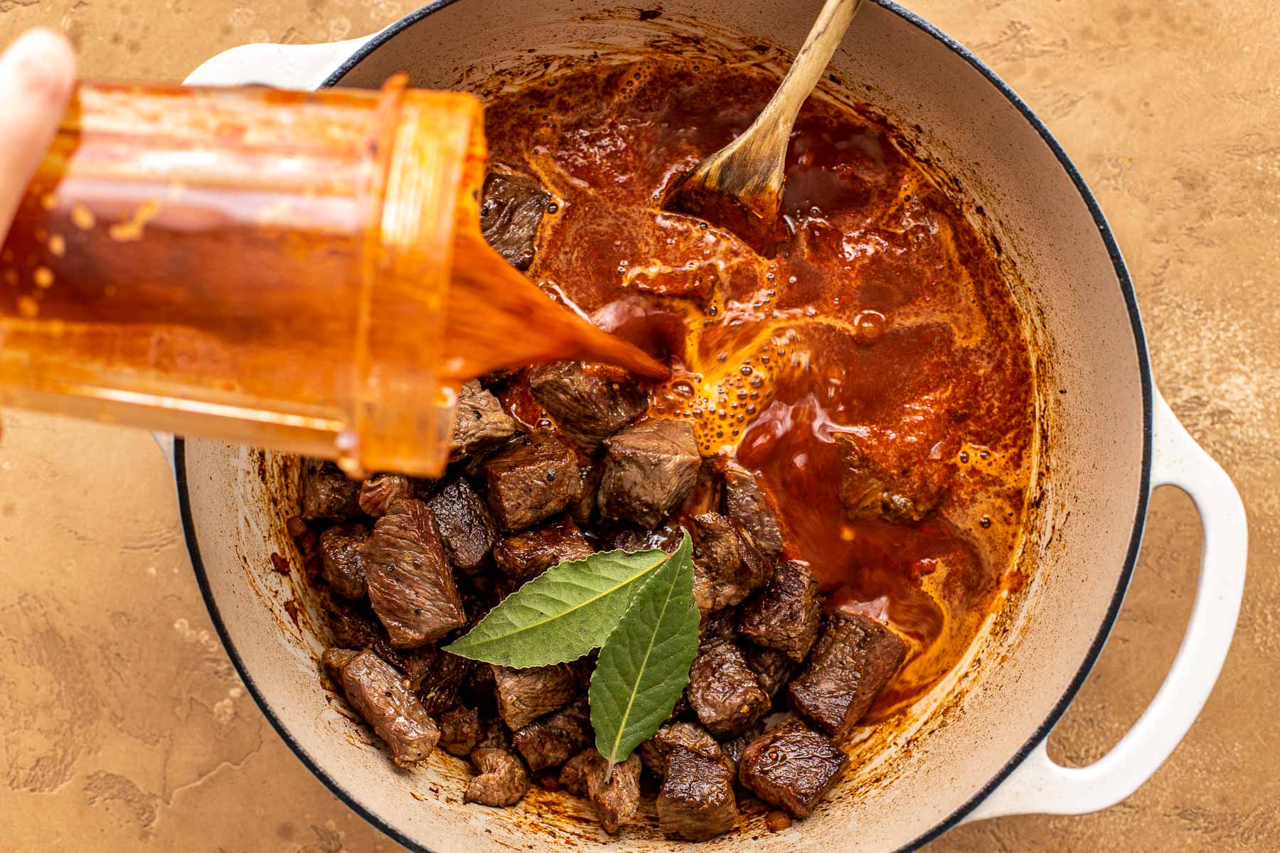 Chunks of beef and bay leaves in a pot as a blended chile sauce is poured in, with a wooden spoon beside.