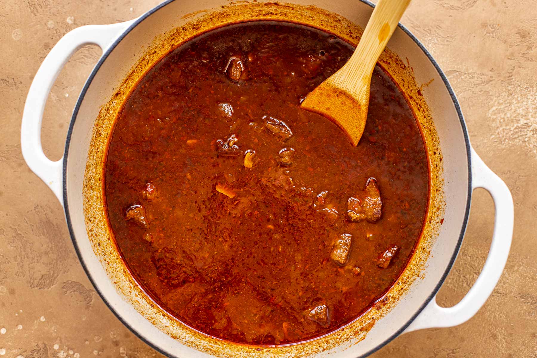 A large white pot filled with a red chile sauce and chunks of beef, with a wooden spoon set in it.