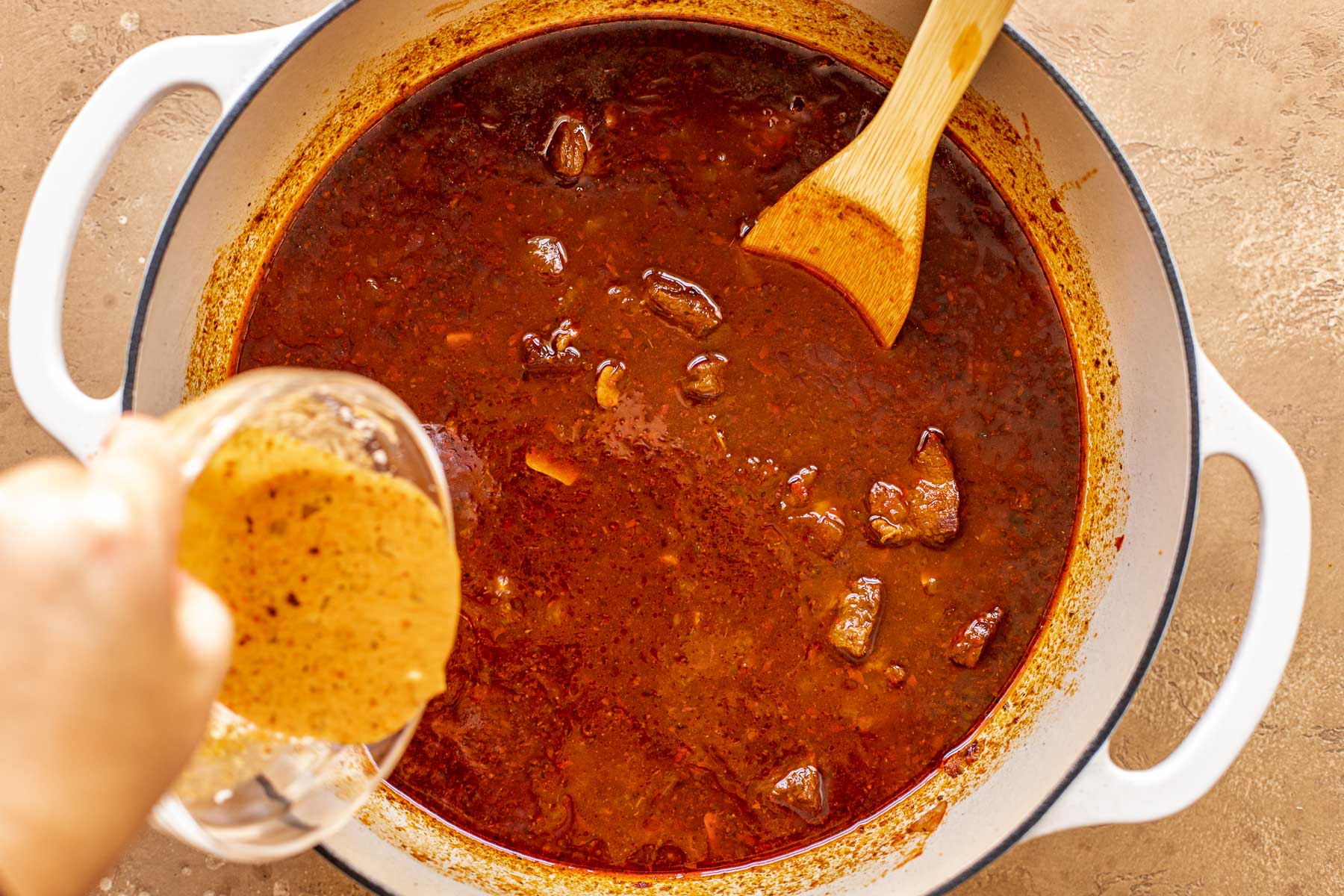 A hand pours a cornstarch slurry into a pot of simmering red stew with chunks of beef and a wooden spoon set inside it.
