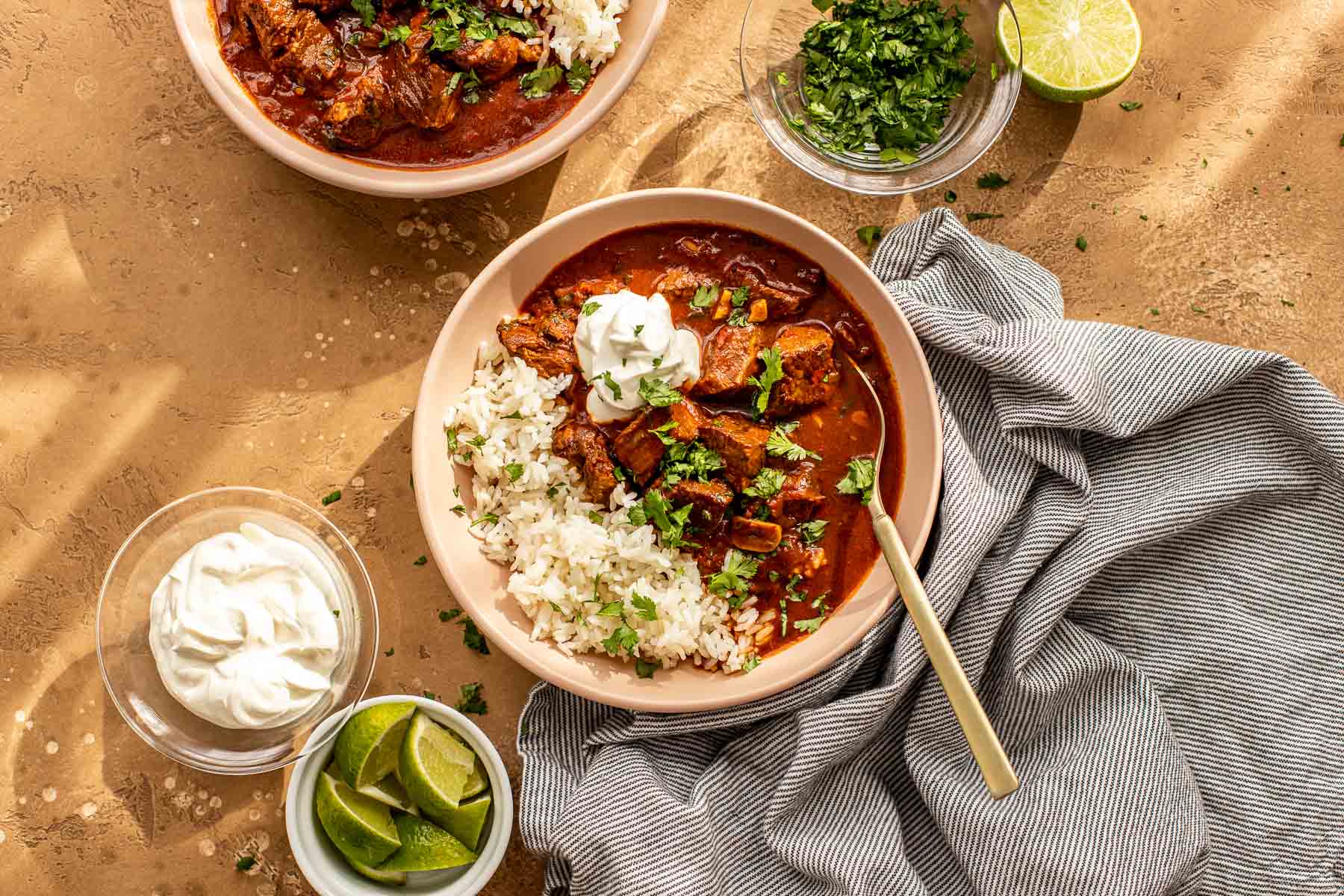 Bowl of chile colorado with rice, topped with sour cream and cilantro, surrounded by lime wedges and more toppings.