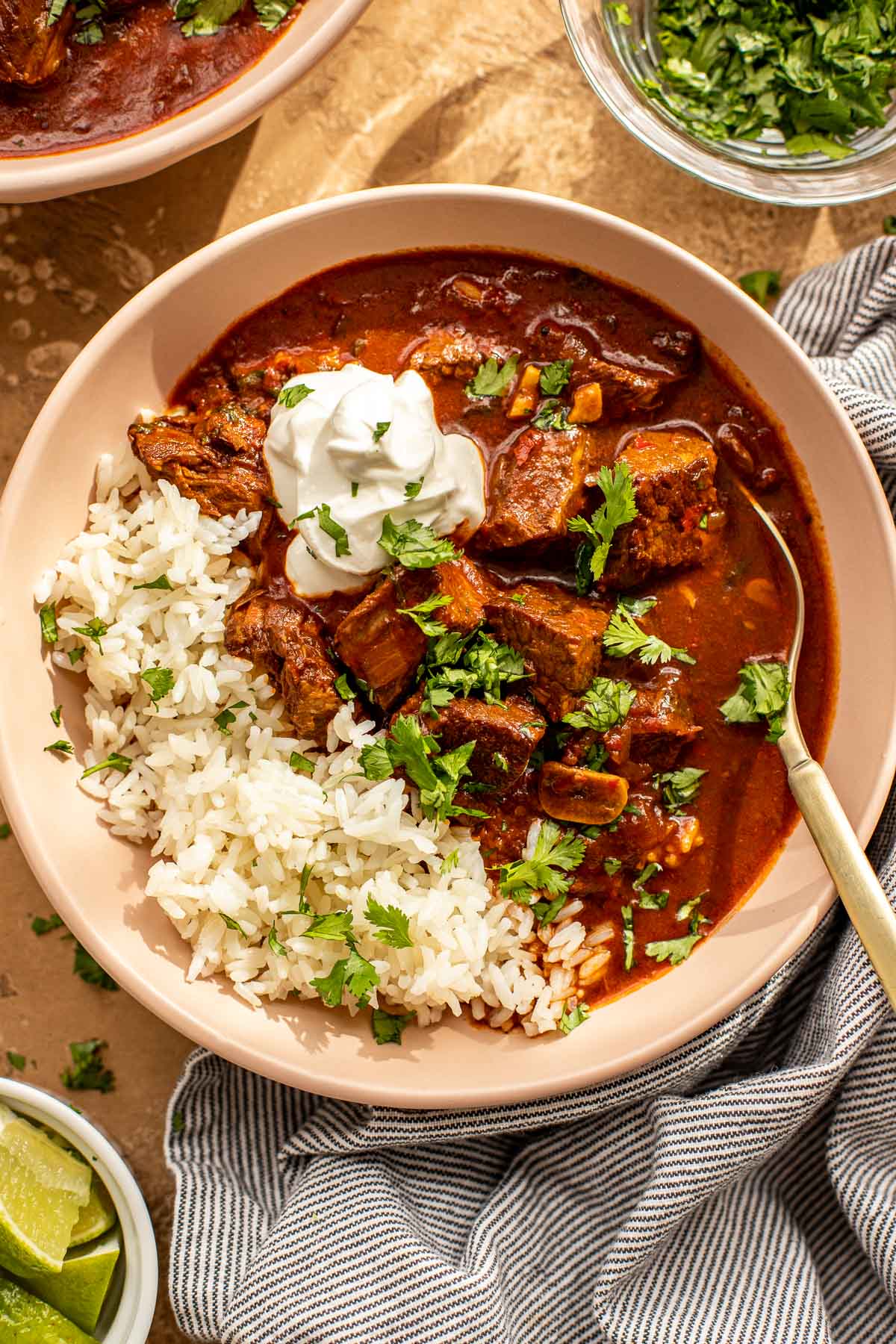 A bowl of rice and beef stew topped with sour cream and chopped cilantro, with lime wedges on the side.