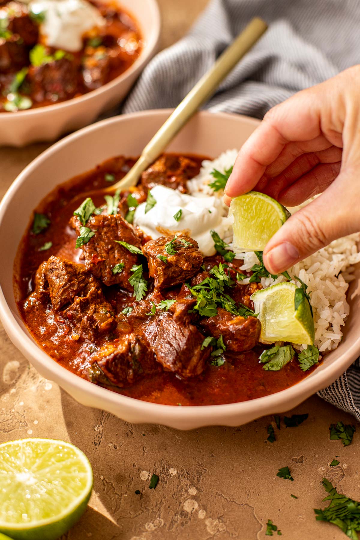 A hand squeezes a lime over a bowl of beef stew with rice, sour cream, and fresh cilantro.