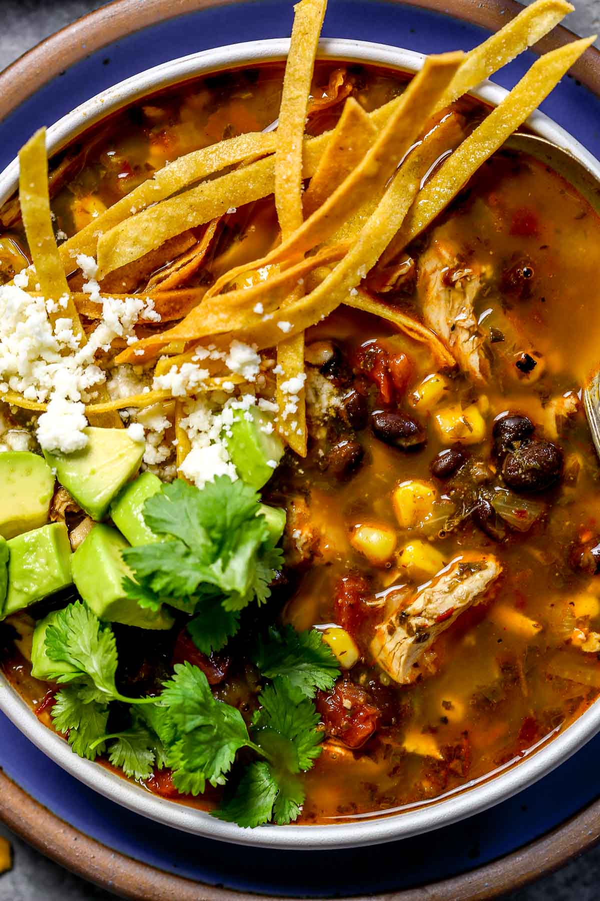 A bowl of chicken tortilla soup topped with avocado, cilantro, tortilla strips, and crumbled Cotijacheese.