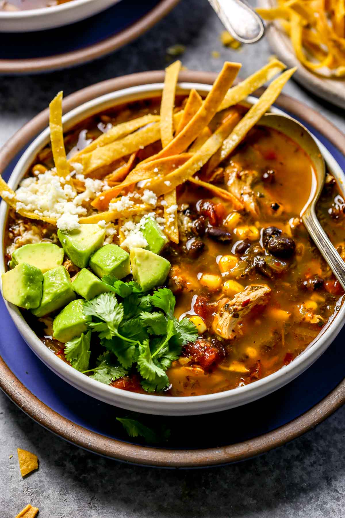 A bowl of chicken tortilla soup topped with avocado, cilantro, tortilla strips, and crumbled Cotijacheese.