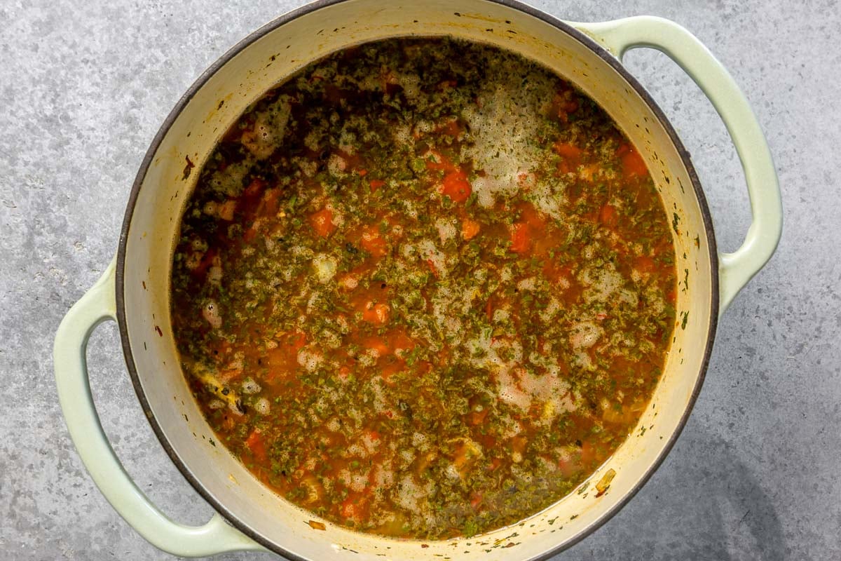 A pot of simmering soup with herbs, tomatoes, and broth on a gray countertop.