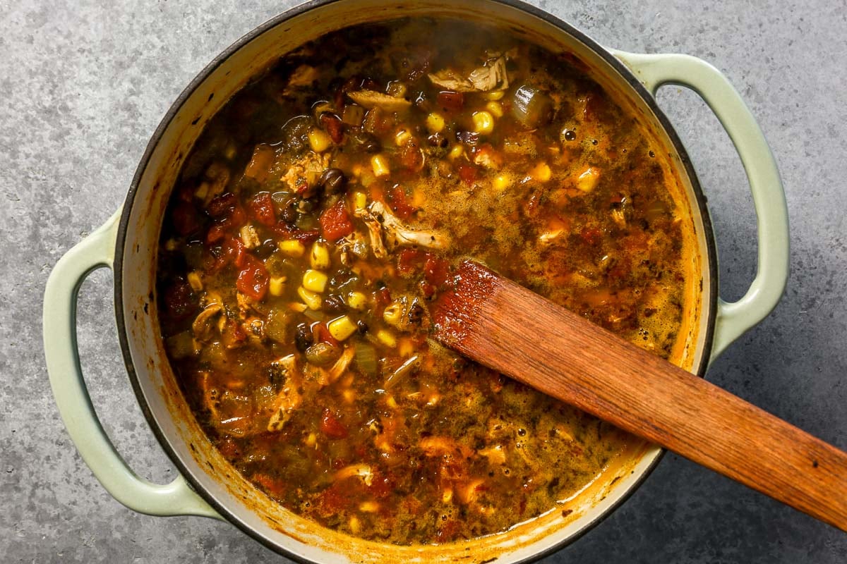 A pot of chicken tortilla soup with corn, tomatoes, beans, and shredded chicken, being stirred with a wooden spoon.