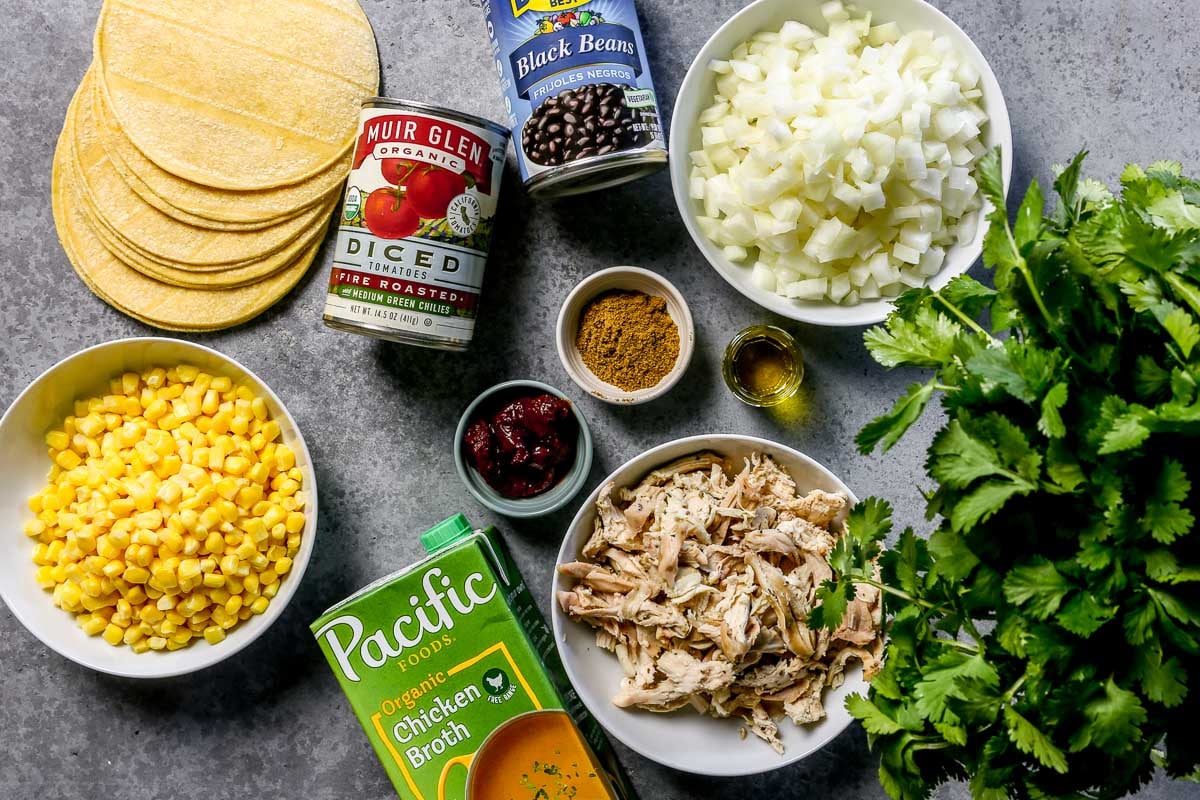 Ingredients for chicken tortilla soup laid out on a counter, including frozen corn, corn tortillas, cooked chicken, broth, black beans, canned tomatoes, diced onion, cilantro, spices, oil, chipotle.