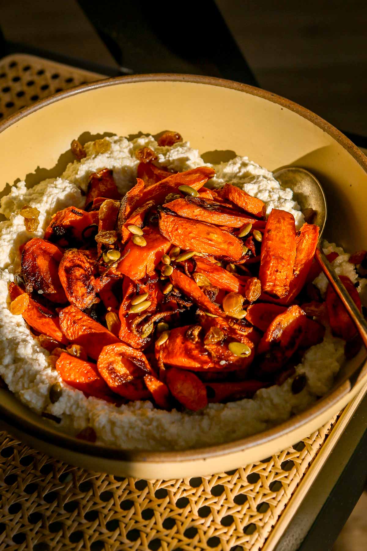 A bowl of whipped farmers cheese topped with roasted carrots, pepitas, and golden raisins, with a spoon on the side.
