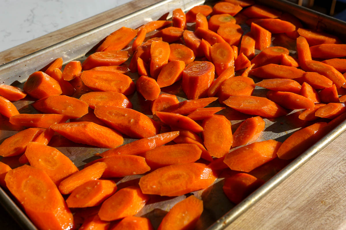 Sliced carrots spread on a baking sheet, ready for roasting, with sunlight shining on them.