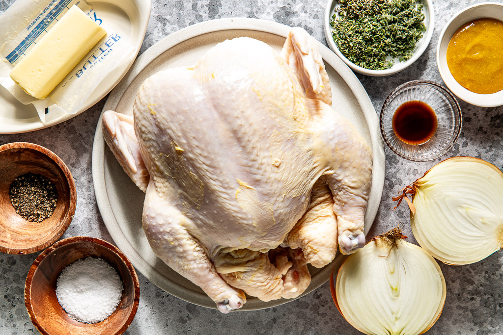 Raw whole chicken on a plate surrounded by butter, herbs, bouillon paste, onion, salt, pepper, and worcestershire sauce.