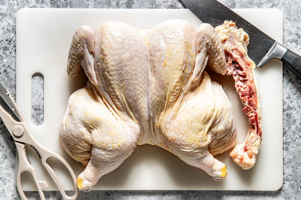 Raw spatchcocked chicken on a cutting board with backbone removed, next to kitchen shears and a knife.