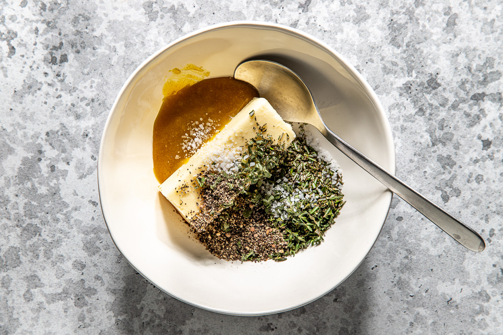 Butter, herbs, salt, pepper, and bouillon paste in a white bowl with a spoon on a speckled gray surface.