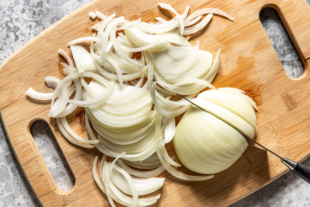 Half-sliced white onion being thinly sliced on a wooden cutting board with a knife.