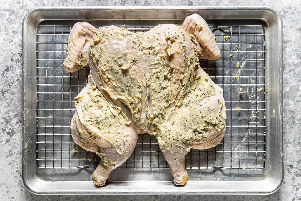Raw spatchcocked chicken coated with herb butter on a wire rack over a baking sheet.