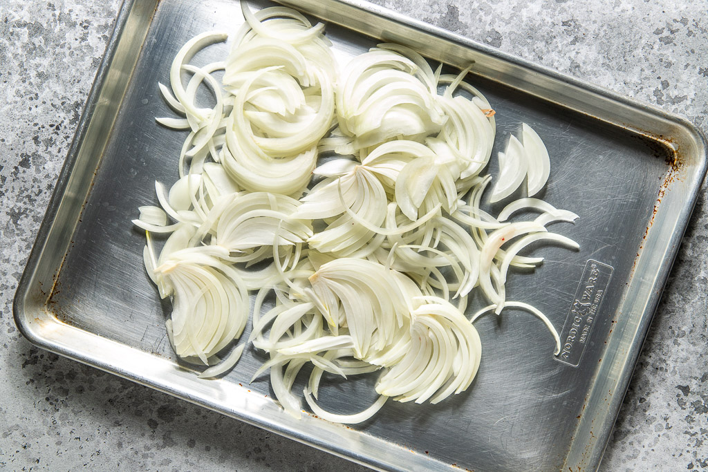 Thinly sliced white onions spread out on a metal baking sheet.